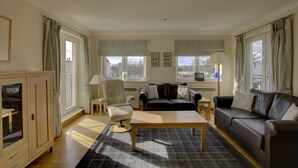 A living room with a sofa, armchair, and coffee table. There are windows with curtains, a lamp, and a cabinet. - Top-floor, two-bedroom golf apartment with views over the famous James Braid Brora Golf Course (Brora)