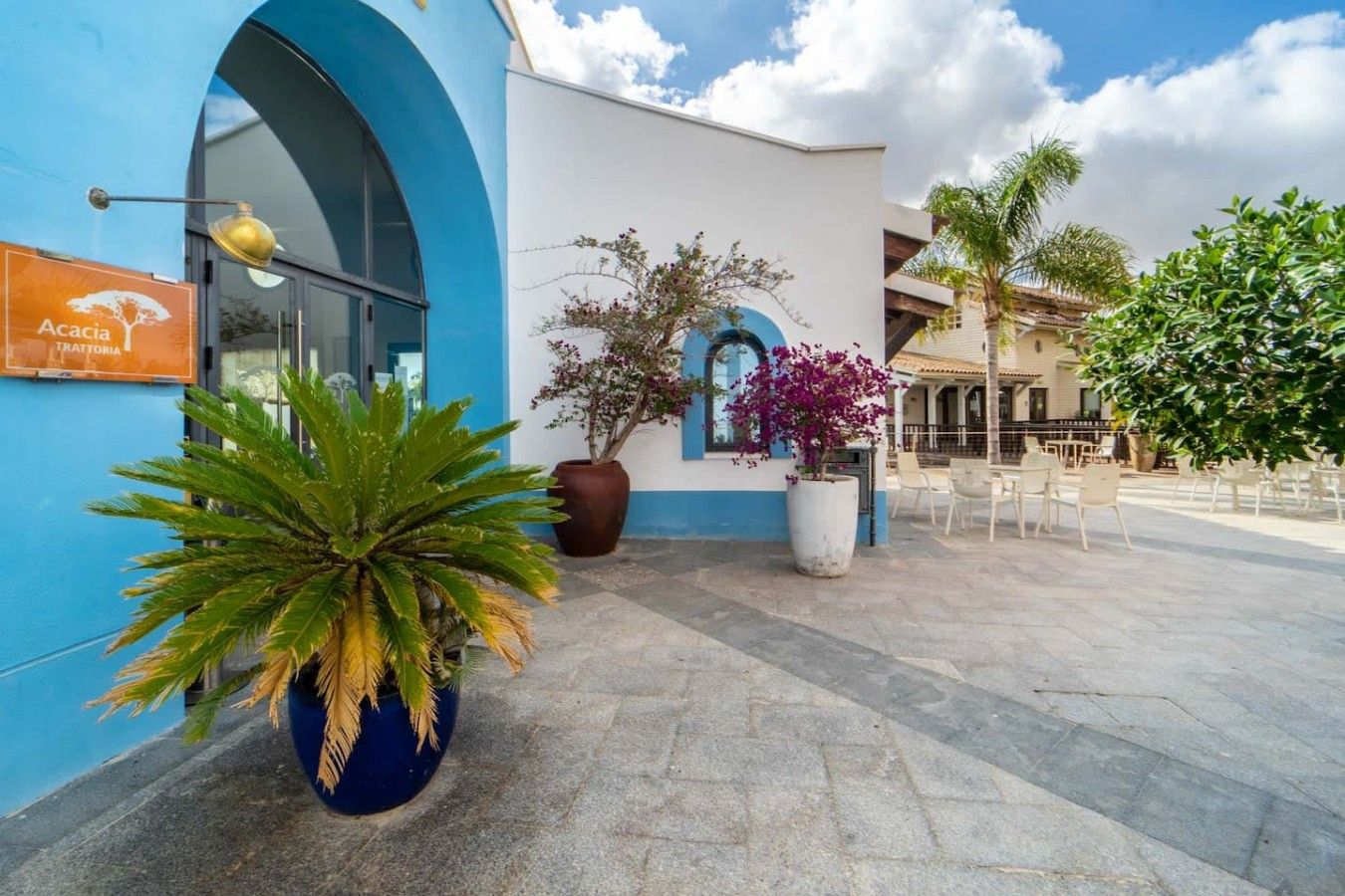 A building with a blue facade, a sign reading 'Acacia Trattoria,' and outdoor seating with chairs and tables.