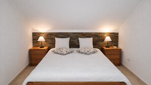 A bedroom with a wooden bed, two bedside tables with lamps, a stone-patterned headboard, and a white wall. - T0 Condomínio jardim da Oura, A/c, piscinas, terra (Albufeira)