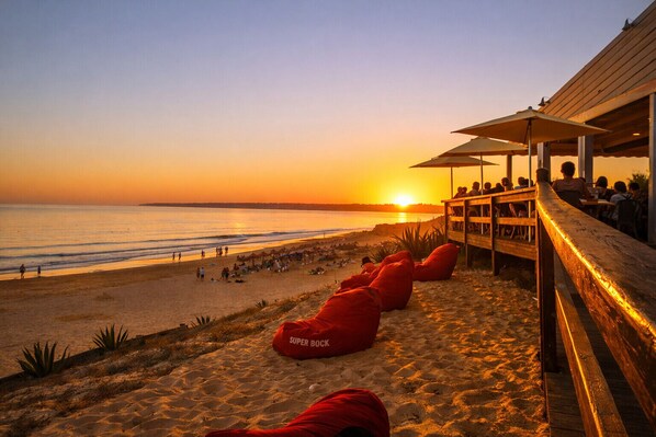 Une vue sur la plage avec une terrasse en bois, des poufs rouges et un coucher de soleil sur l’océan.