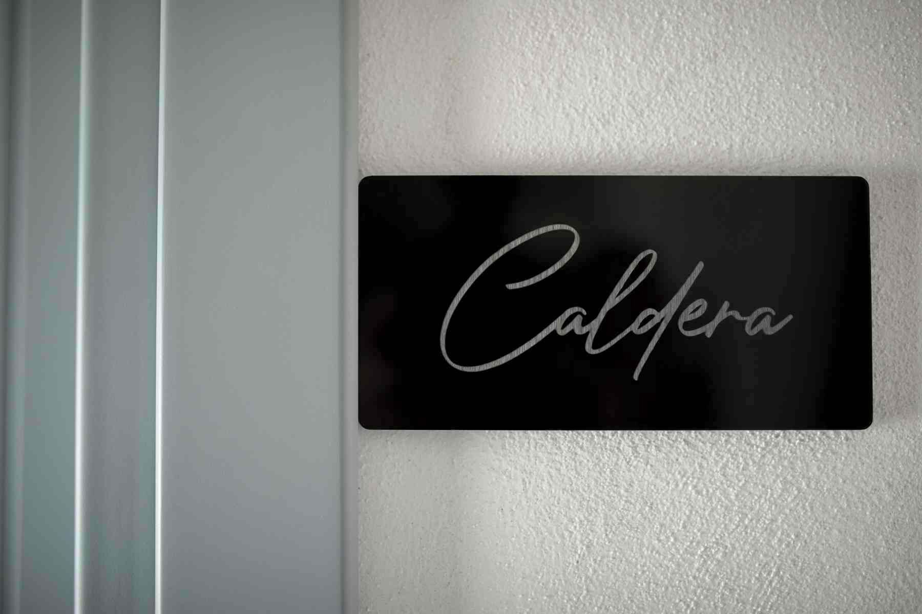 Ein schwarzes Schild mit der Schriftart 'Caldera' in verspielten Buchstaben an einer weißen Wand.