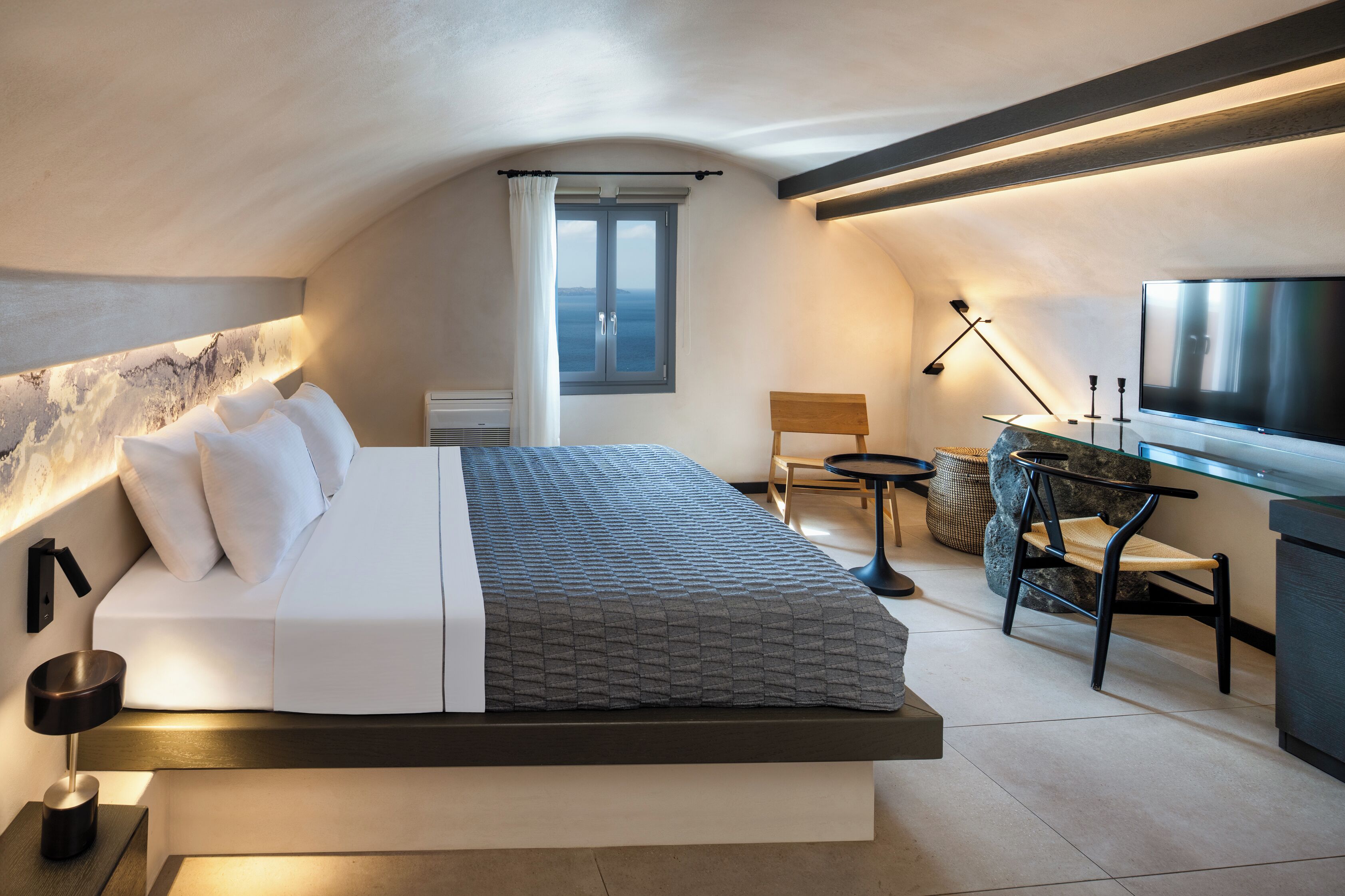 Une chambre à coucher moderne, avec un lit, une table de chevet, une lampe et une vue sur la mer par la fenêtre.