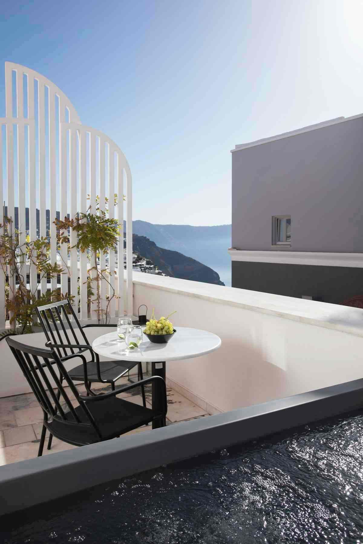En balkon med et rundt bord, to stole, en skål med frugt og havudsigt.