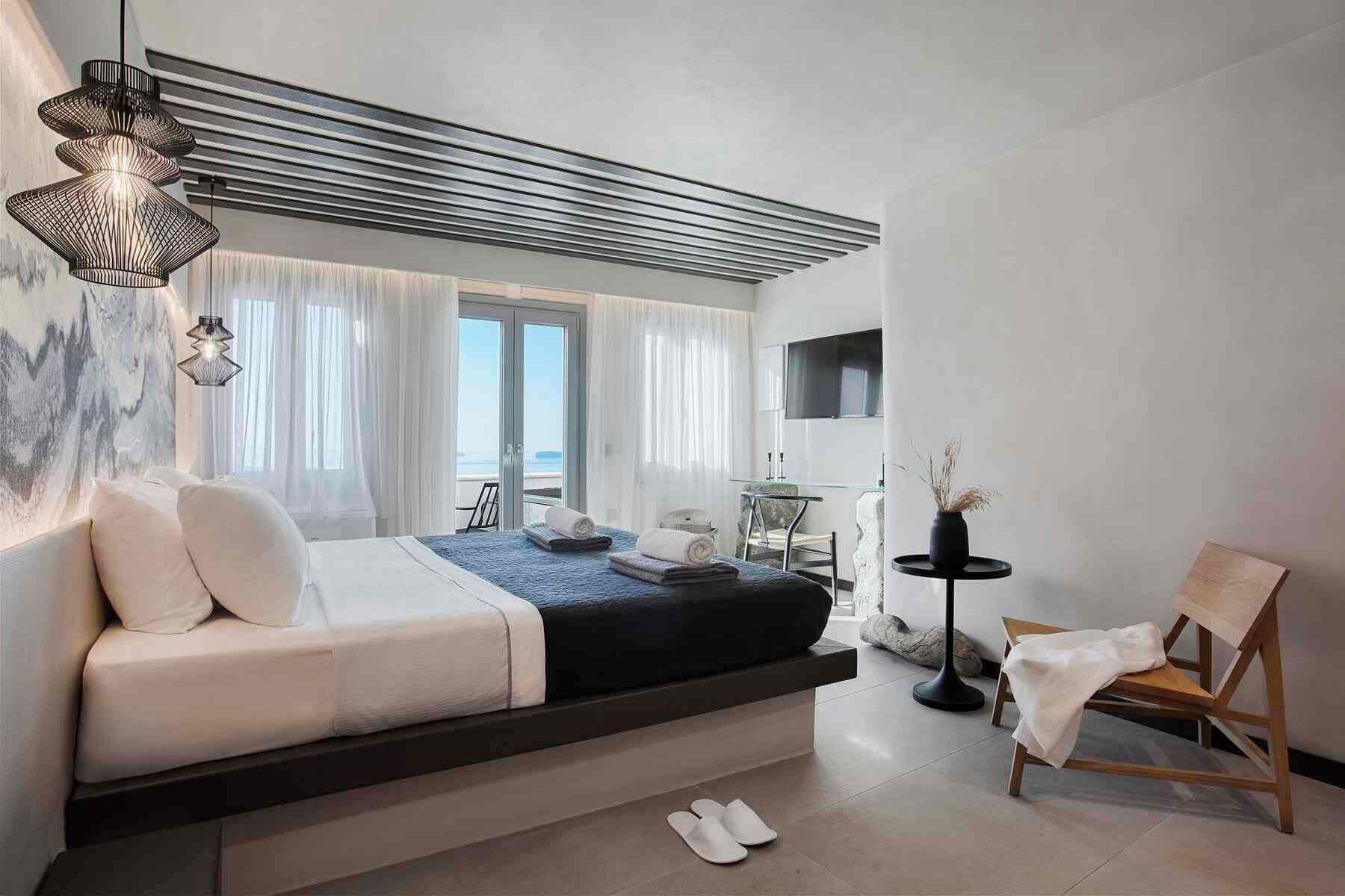 A modern hotel room with a bed, a seating area, a TV, and a view of the sea.