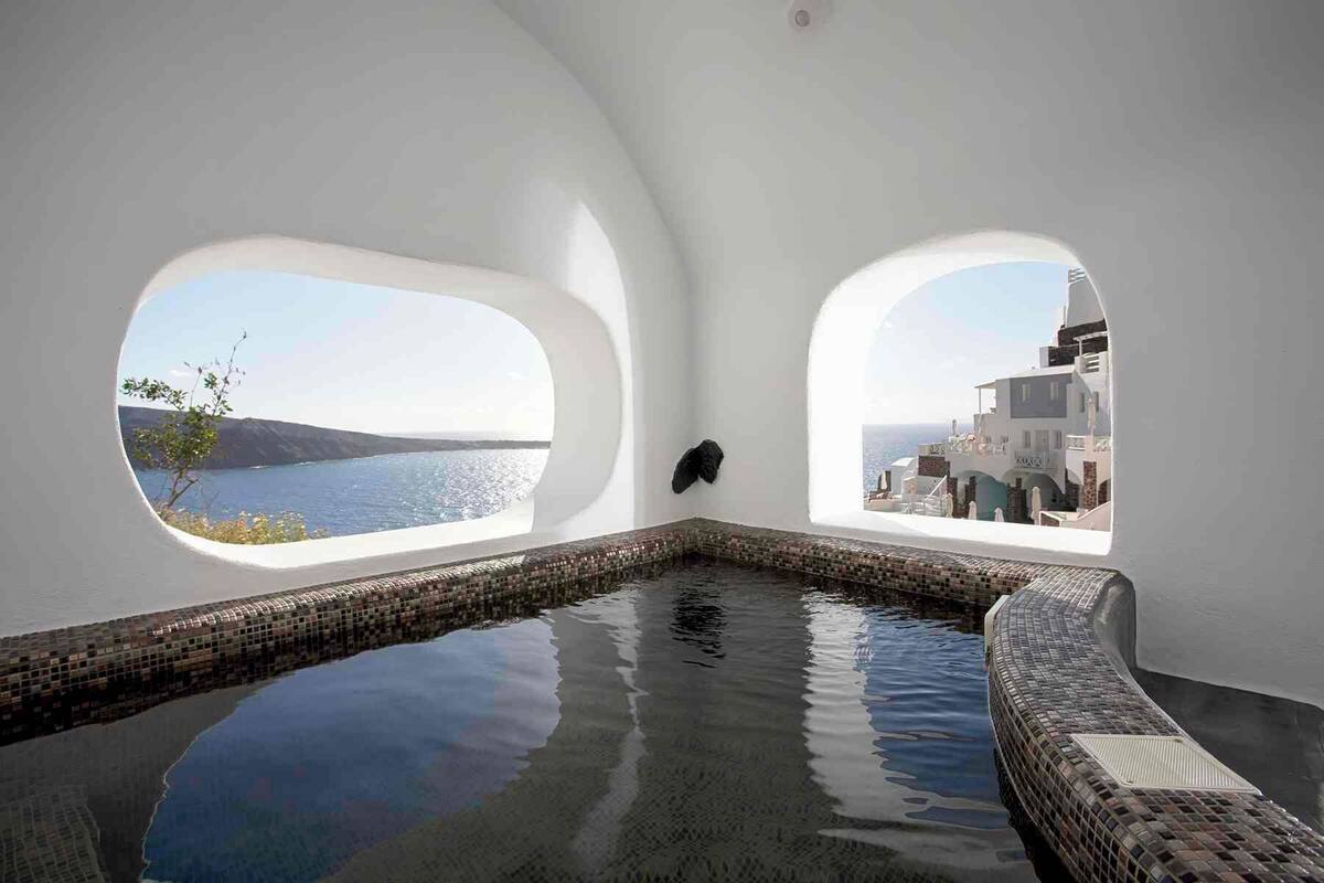 Une piscine intérieure moderne, dotée de deux grandes fenêtres en arc qui offrent une vue sur la mer et un bâtiment blanc.