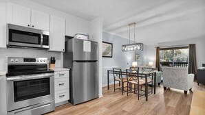 A modern kitchen with stainless steel appliances, a dining area with a table and chairs, and a view of the outdoors through a window. - Tennis Villa D2 - 2BR Walk to Beach & Pool (Hilton Head Island)