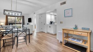 A modern living room with a dining area, a wooden desk, and a view through a large window. - Tennis Villa D2 - 2BR Walk to Beach & Pool (Hilton Head Island)