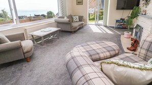 Ein Wohnzimmer mit Sofa, Sessel und Couchtisch. Ein Fernseher ist an der Wand befestigt, und ein Fenster bietet Blick ins Freie.