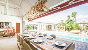 A dining area with a long wooden table set for a meal, overlooking a pool with lounge chairs and palm trees. - Swiss 2 Villa in Dalung (Tibubeneng)