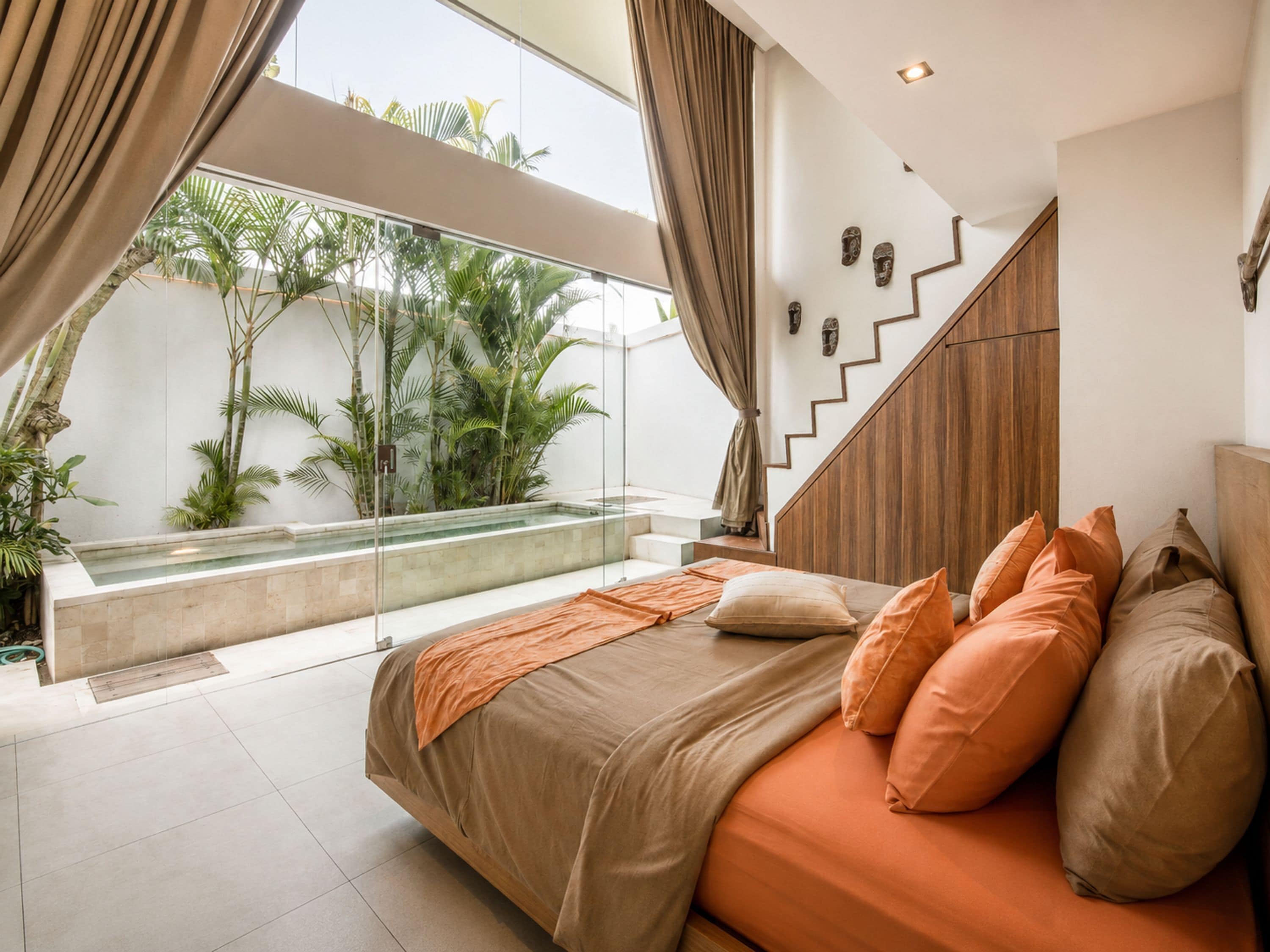 A bedroom with a bed, pillows, a staircase, and a view of a pool and plants.