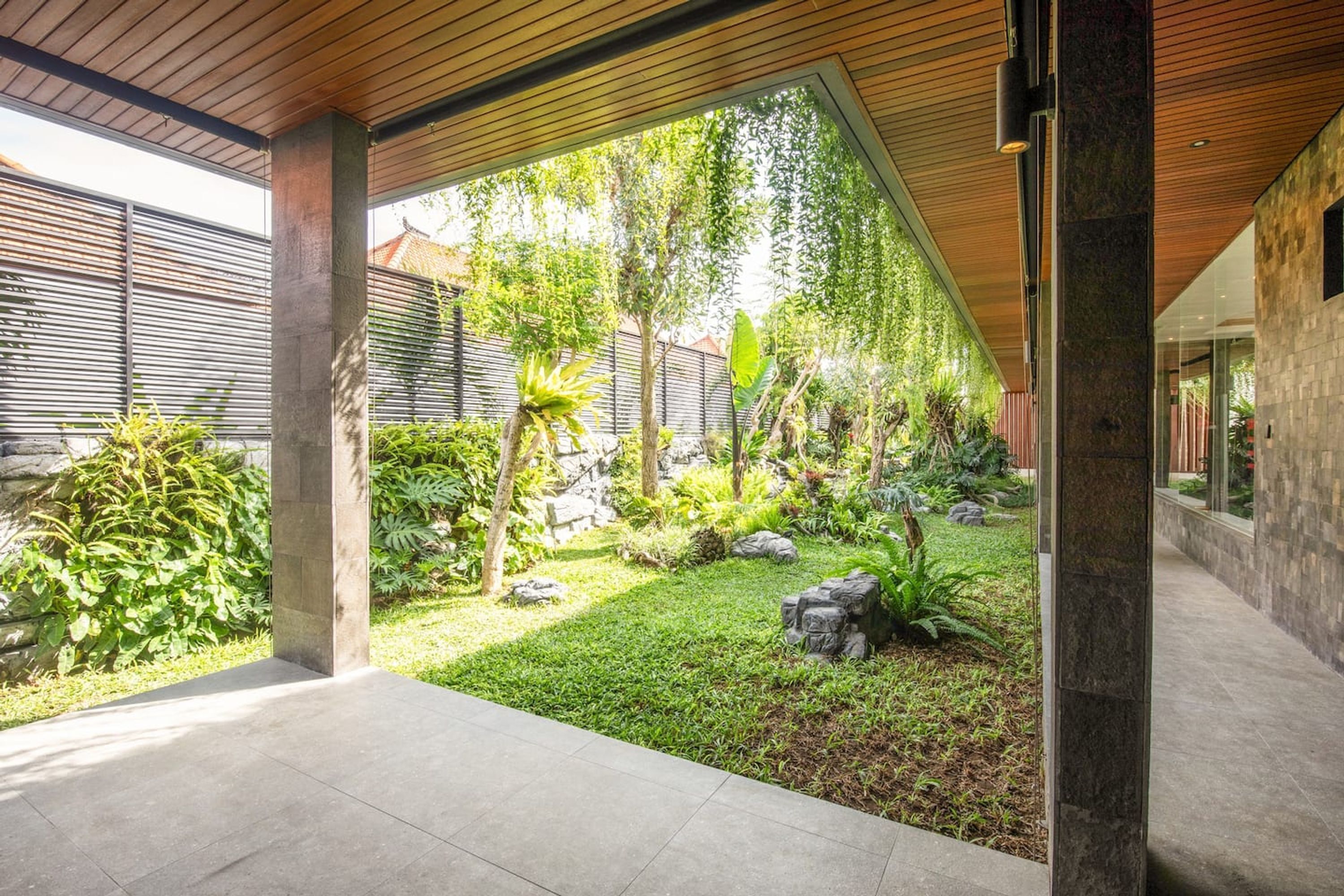 Un patio exterior moderno con techo de madera, suelo de hormigón y una variedad de plantas y rocas.