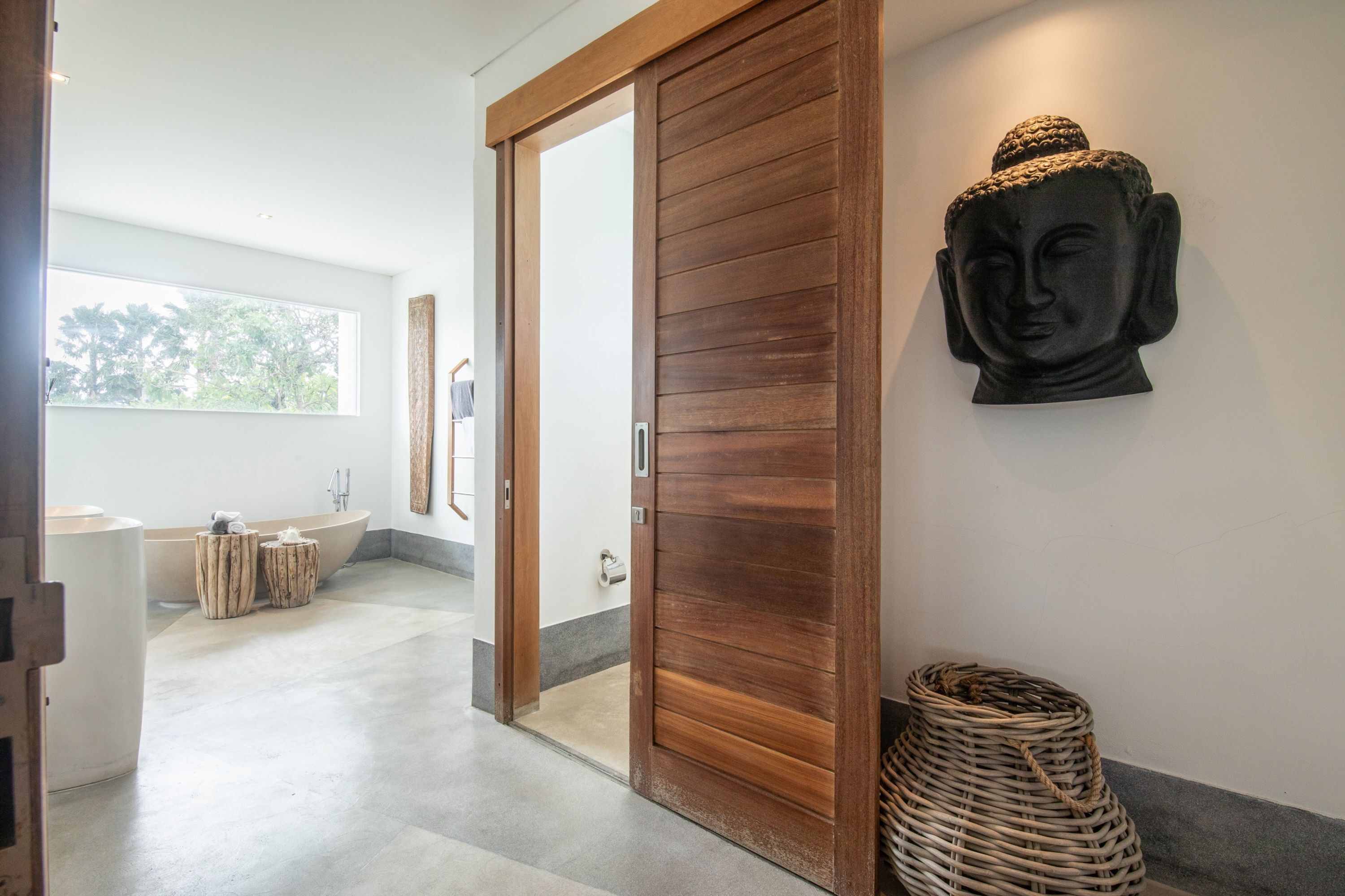 A modern bathroom with a large bathtub, a wooden door, and a Buddha head sculpture on the wall.