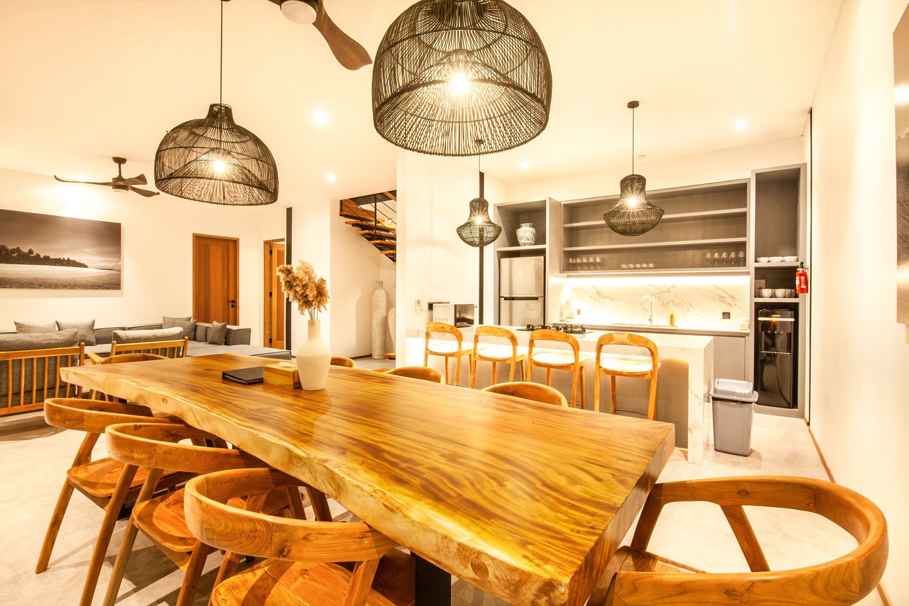 Un espace de salle à manger moderne avec une grande table et des chaises en bois, des luminaires suspendus et une cuisine en arrière-plan.