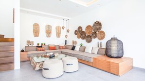 Een moderne woonkamer met een hoekbank, poefjes, een glazen salontafel en decoratieve wandkunst.