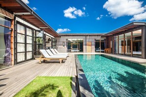 A modern outdoor pool area with wooden deck, lounge chairs, and glass doors leading to a covered patio. - Bodhisatva in Pererenan (Pererenan)
