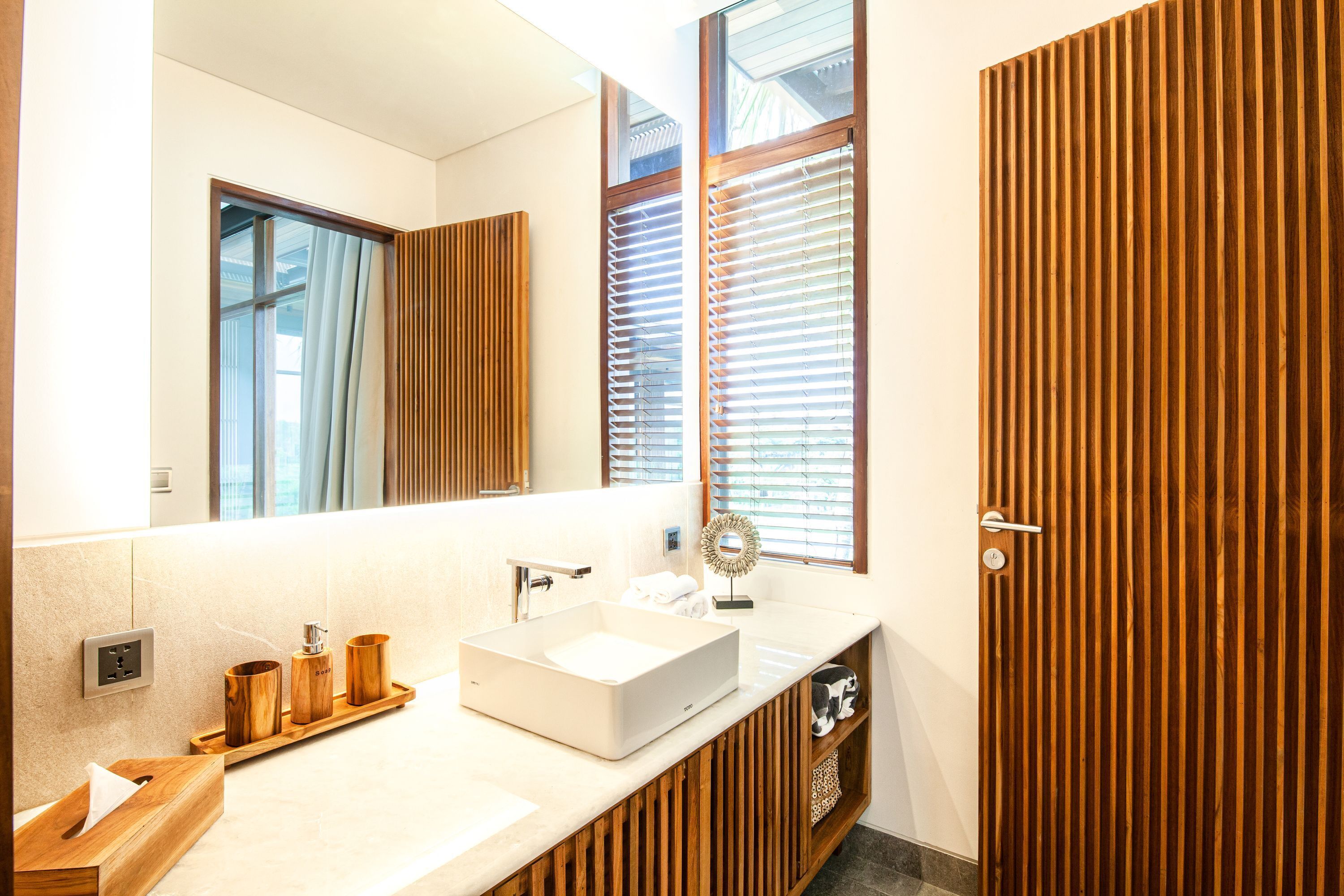 Un baño moderno con un espejo grande, paneles de madera, un lavabo blanco y una ventana con persianas.