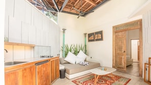 A compact kitchen with wooden cabinets, a built-in dishwasher, and a sink. There's a beige sofa with pillows, a small round table, and a patterned rug. A large plant is in the corner, and a wooden door leads to another room. - BA Villa 5 1 Bed Villa 5 Mins to Every Exquisite 1-br Villa With Pool 4 Mins to Echo Be (Canggu)