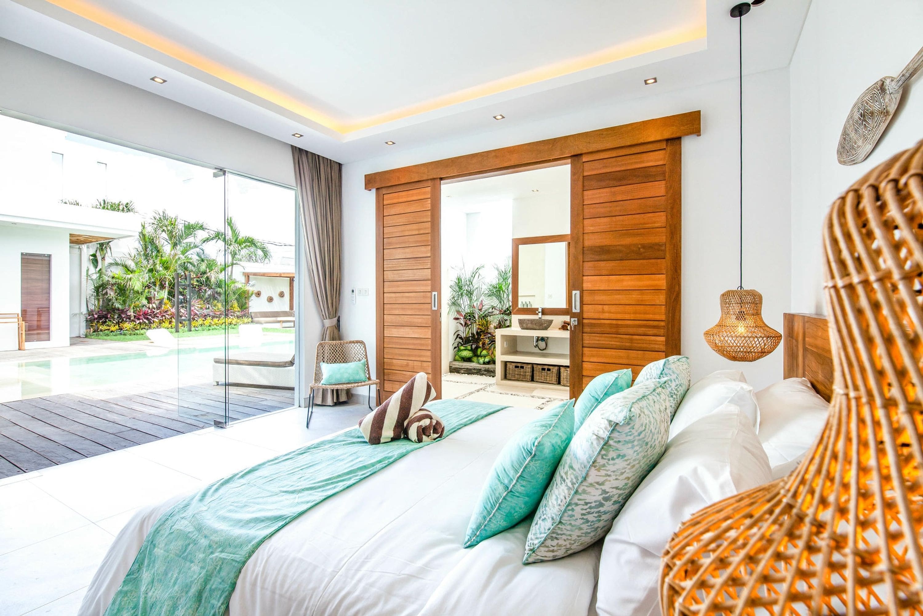 Une chambre moderne avec un grand lit, une tête de lit en bois, une porte en verre donnant sur un balcon et une vue sur un jardin.