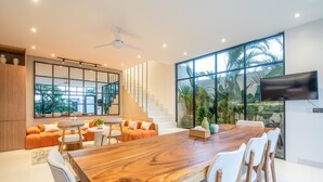 A spacious living area with a large wooden dining table, white chairs, and a sofa set. A staircase leads to an upper level, and a TV is mounted on the wall. - Mateo Villa Marvelous 6BR 2 Mins to Beach (Tibubeneng)