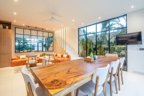 A spacious living area with a large wooden dining table, white chairs, and a sofa set. A staircase leads to an upper level, and a TV is mounted on the wall. - Mateo Villa Marvelous 6BR 2 Mins to Beach (Canggu)