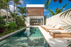 Un área de piscina moderna con sillones de descanso y una piscina cristalina, rodeada de plantas tropicales.