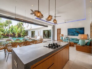 A modern kitchen with a central island, dining area with wicker chairs, and a living room with a sofa and a painting. - Marina Villa in Seminyak (Seminyak)