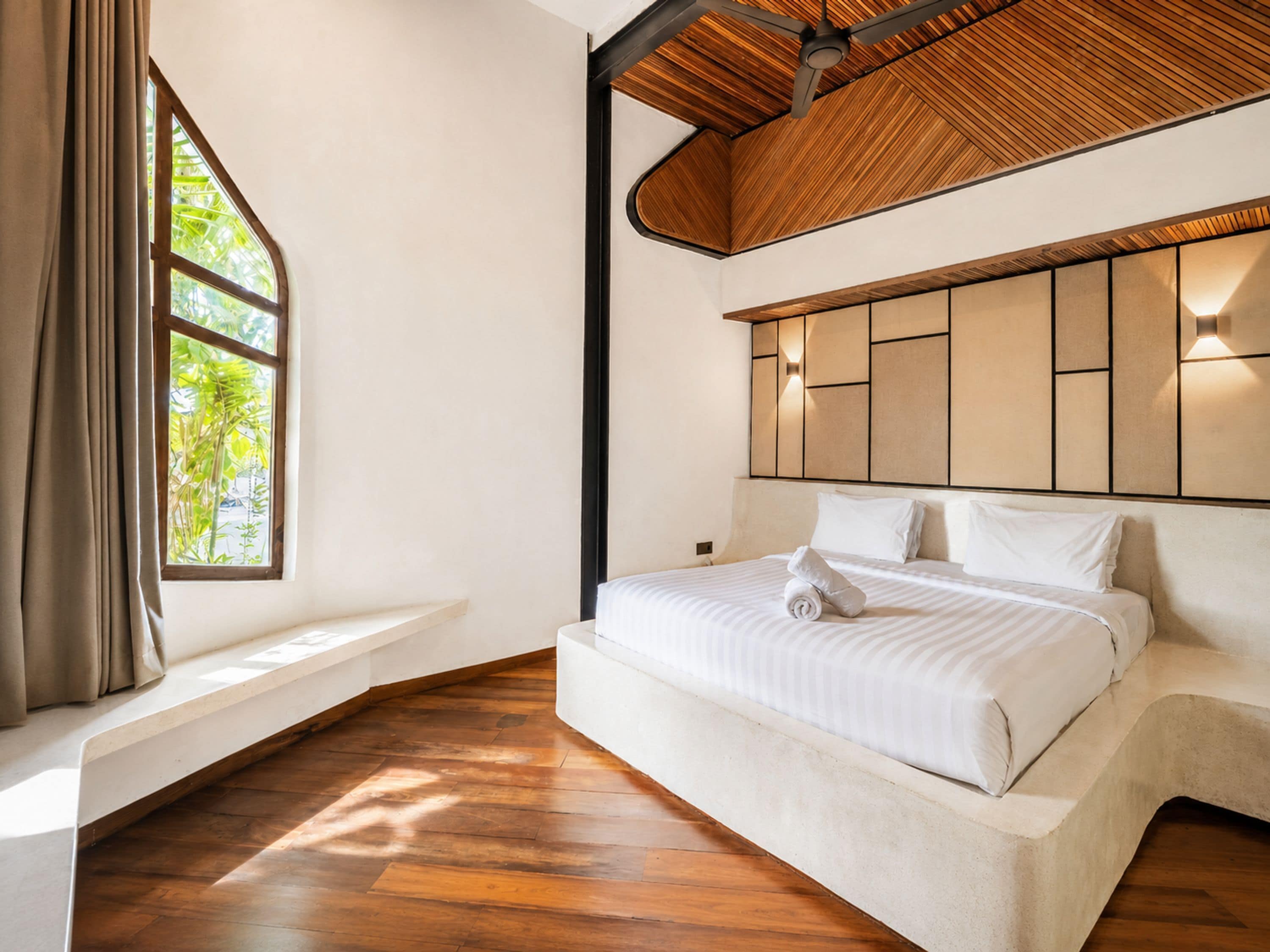 A bedroom with a large bed, a window with curtains, a wooden ceiling, and a wall-mounted light fixture.