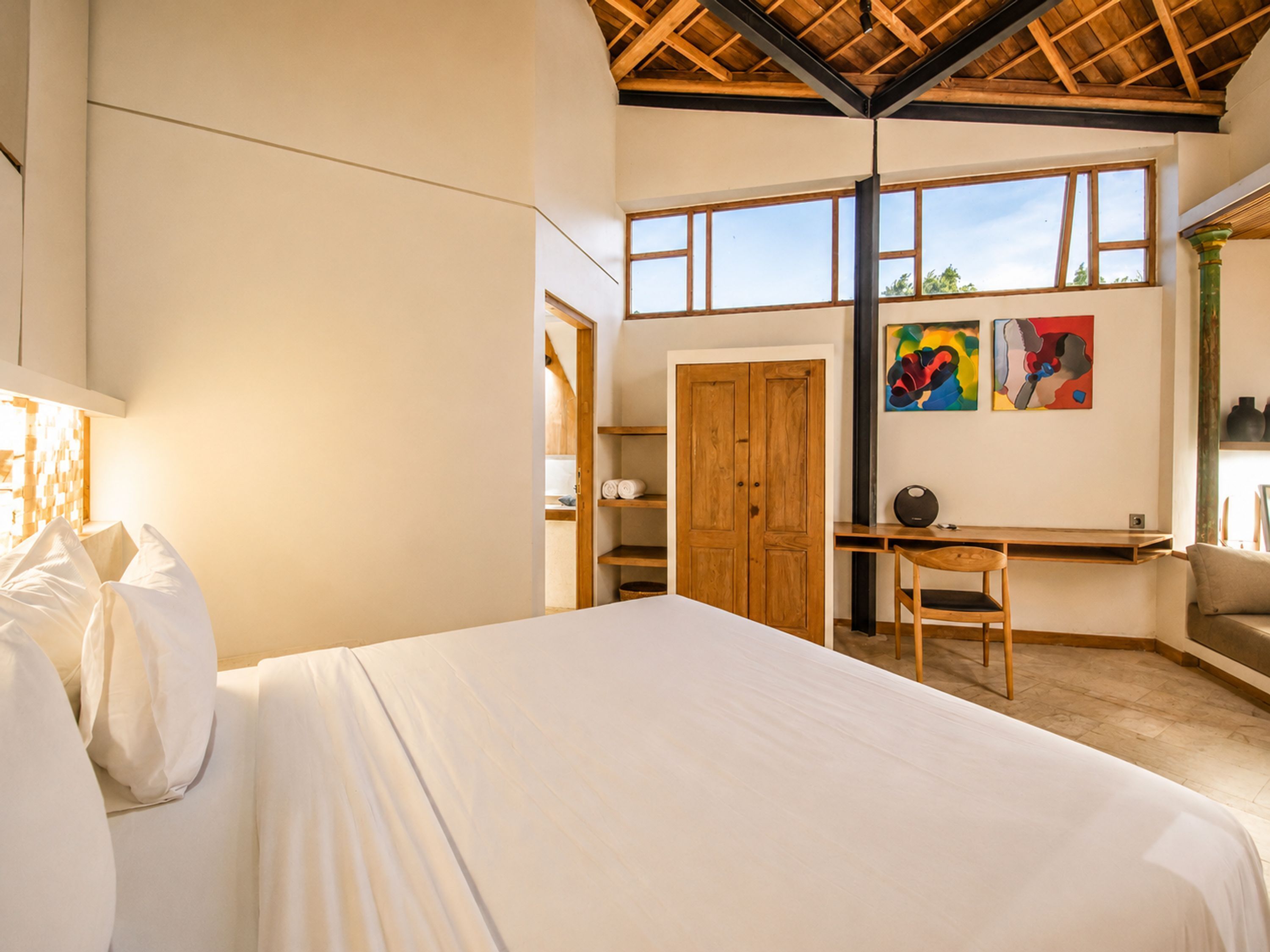 A bedroom with a bed, a desk, a chair, a wardrobe, and a window with artwork.