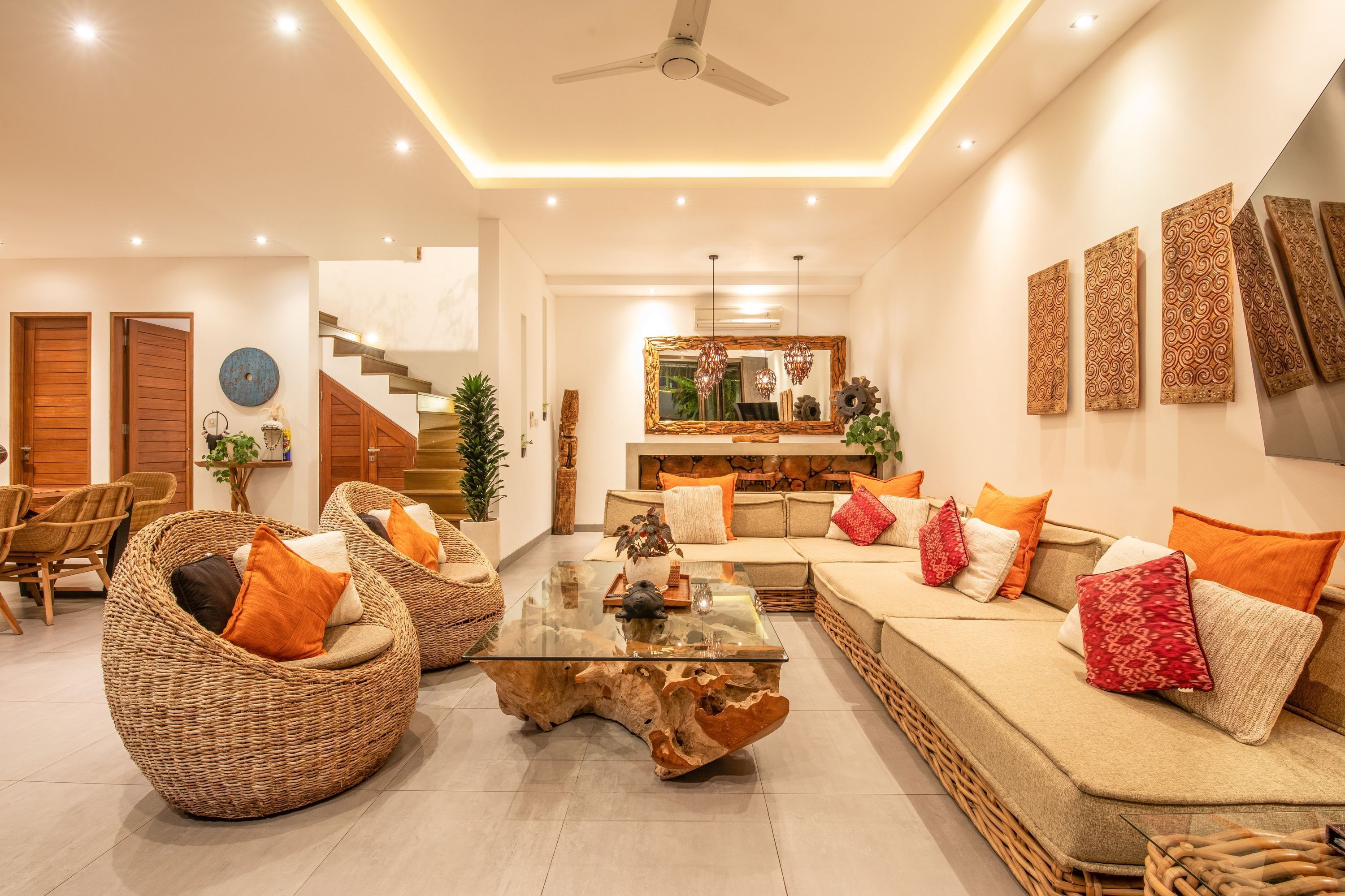A modern living room with a glass coffee table, wicker chairs, a sofa with orange cushions, and a staircase leading to another level.