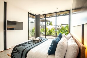A modern bedroom with a large bed, a flat-screen TV, and a view of greenery through large windows.