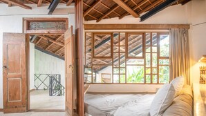 Una habitación con techo de madera, una ventana con vistas a la vegetación, una cama con almohadas y una puerta de madera.