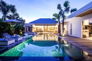 A modern house with a swimming pool, lounge chairs, and a well-lit interior. - Canggu Julia 2 in Dalung (Tibubeneng)