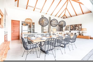 A spacious dining area with a long wooden table and black wire chairs, a kitchen with white cabinets, and a high ceiling with wooden beams. - Canggu Julia 2 in Dalung (Canggu)