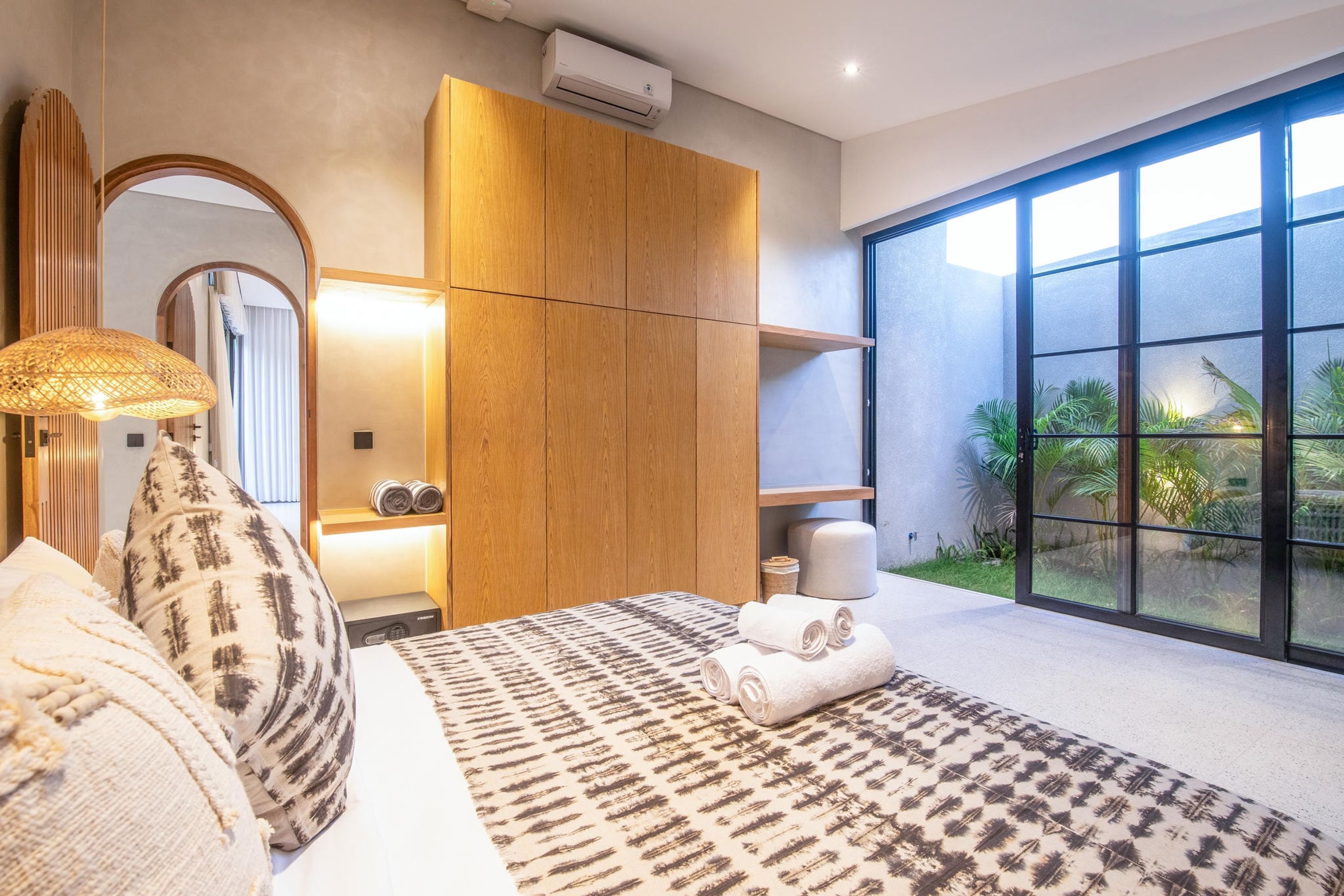 A modern bedroom with a large bed, a wooden wardrobe, a mirror, and a view of greenery through large windows.
