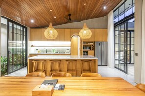 A modern kitchen with wooden cabinets, a central island with bar stools, and pendant lighting. - Villa Serai 5br to the Beach (Pecatu)