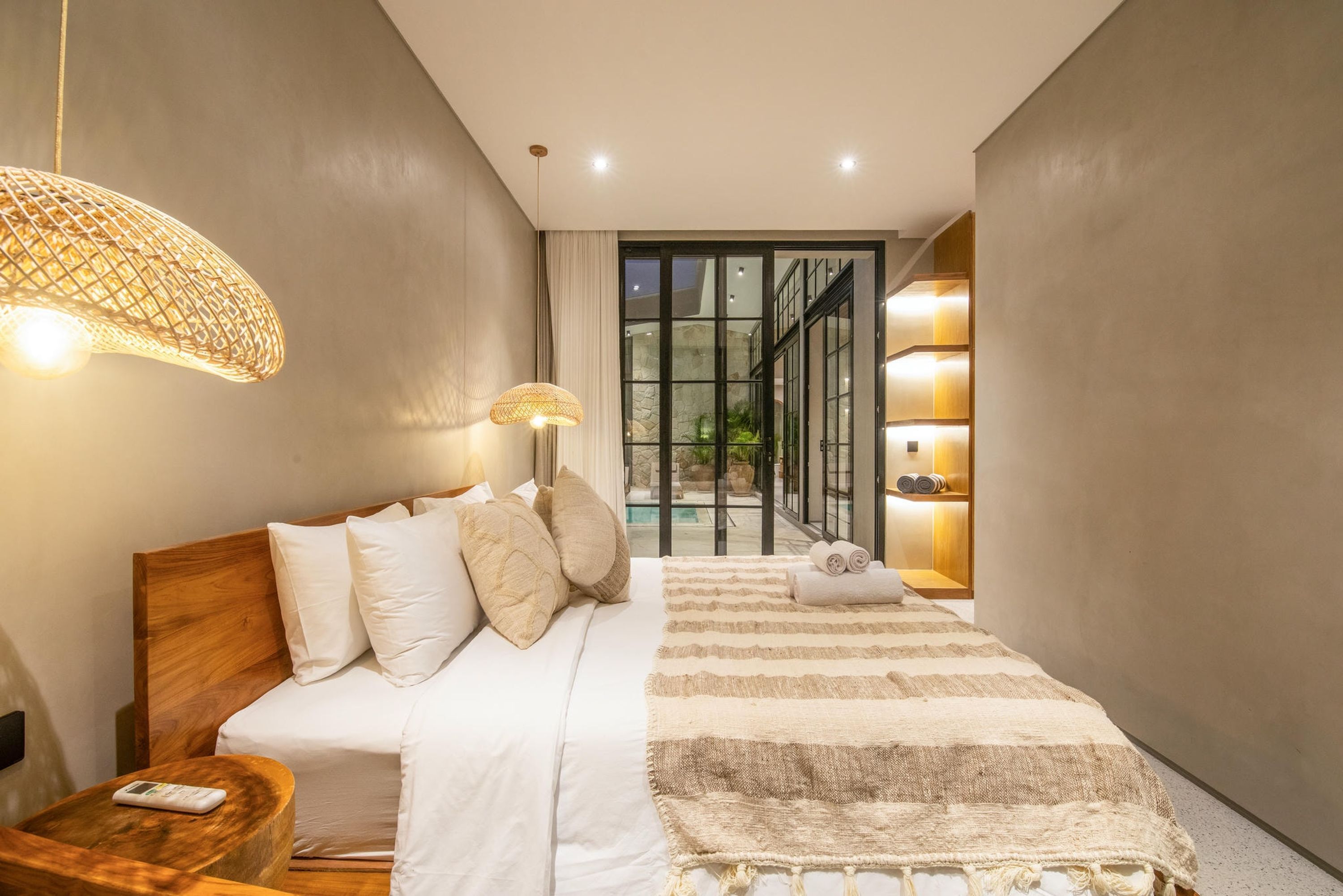 A modern hotel room with a large bed, a small bedside table, a bedside lamp, and a view of a pool through a glass door.
