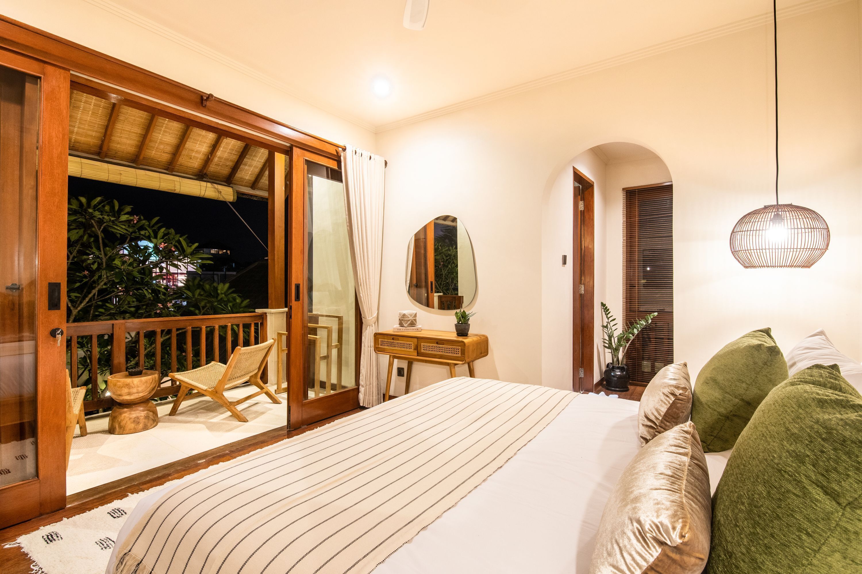 A bedroom with a large bed, a wooden desk, a mirror, and a balcony with a view of the night sky.