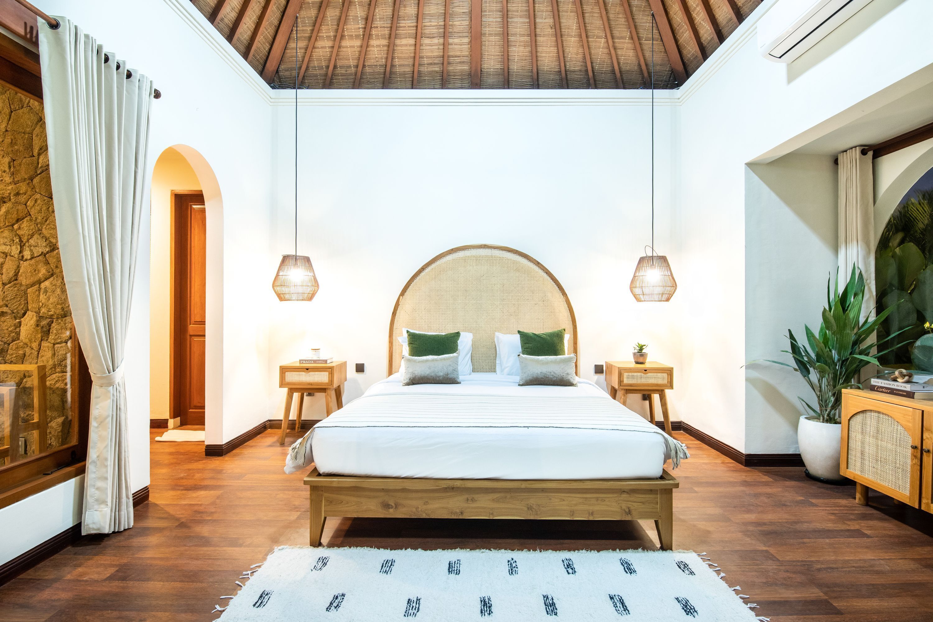 A bedroom with a wooden bed, two bedside tables, a stone wall, a wooden ceiling, and a large window.