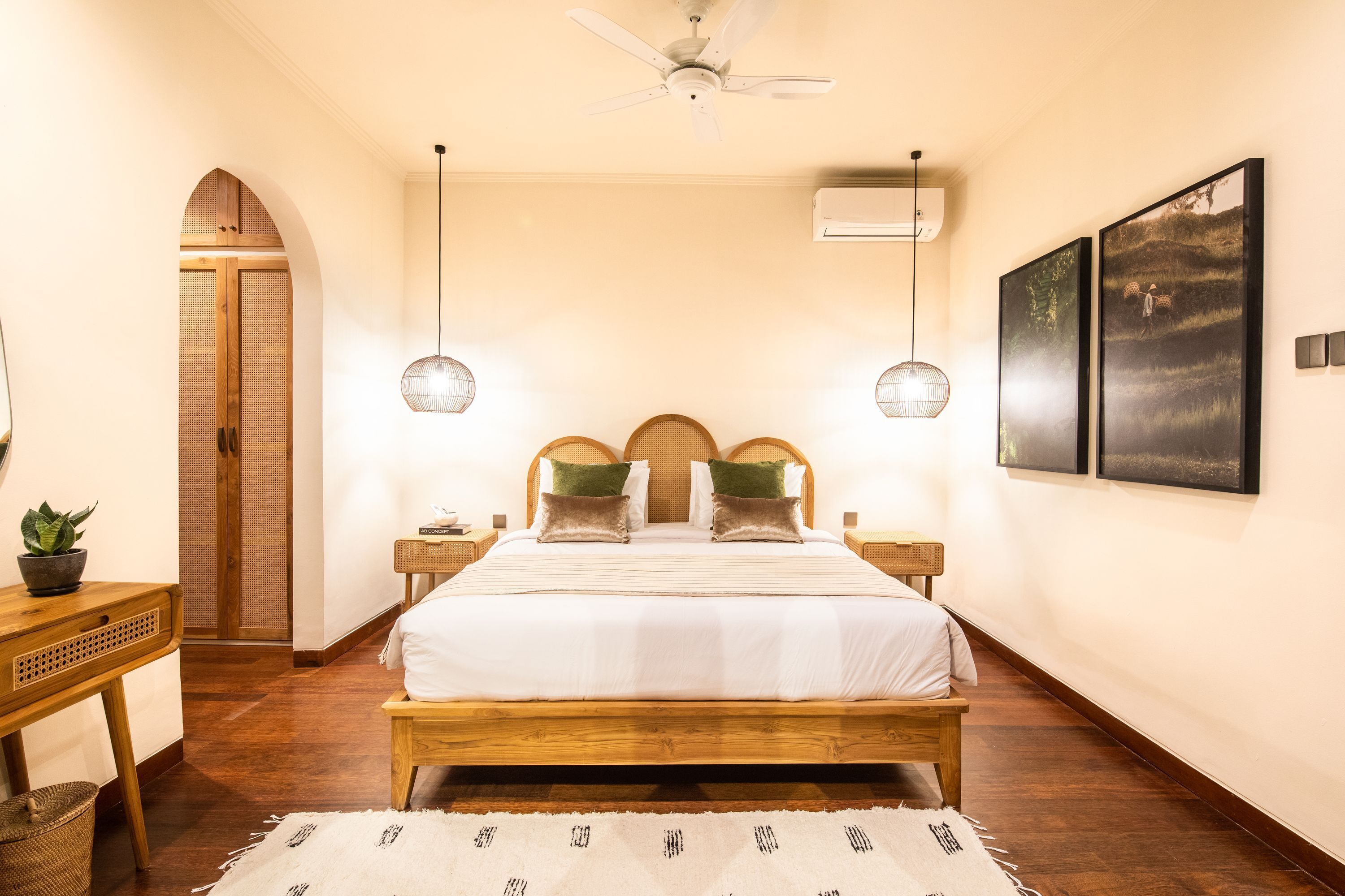 Une chambre à coucher avec un lit en bois, deux tables de chevet, un ventilateur de plafond, un climatiseur mural et des œuvres d’art encadrées.