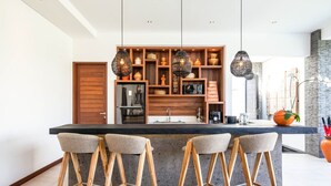Eine moderne Küche mit einer zentralen Kochinsel, Holzhocker, hängenden Pendelleuchten und verschiedenen dekorativen Accessoires auf Regalen.