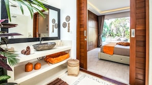 Ein modernes Badezimmer mit einem großen Spiegel, einem Becken aus Stein und einer Holztür, die zu einem Schlafzimmer mit Bett und Blick auf einen Pool führt.