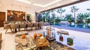 Ein modernes Wohnzimmer mit einem Glastisch, einem Essbereich mit Holztisch und Stühlen sowie Blick auf einen Pool mit Palmen.