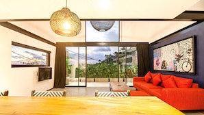 A modern living room with a wooden dining table, red sofa, and large windows offering a view of greenery. - Loft 1 App in Pererenan (Munggu)