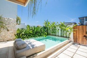 Una piscina con un muro de piedra, abundante vegetación, una terraza de madera y un edificio moderno al fondo.