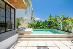 Un área de piscina exterior moderna con una pared de piedra, abundante vegetación y una zona de estar con cojines.