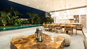 Un área de comedor al aire libre con piscina, mobiliario de madera e iluminación colgante.