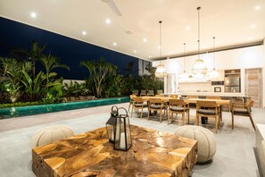 Un área de comedor al aire libre con piscina, mobiliario de madera e iluminación colgante.