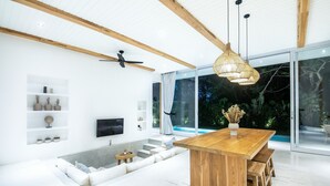 Een moderne woonkamer met een houten eettafel, witte banken, een televisie en een groot raam met uitzicht op een tuin.
