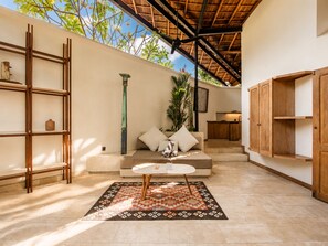 A sunlit interior with a sofa, a small table, a patterned rug, a wooden shelf, and a kitchen area. - BA Villa 4 1BR Lush 1-br Villa With Pool 4 Mins to Echo Beach (Pererenan)