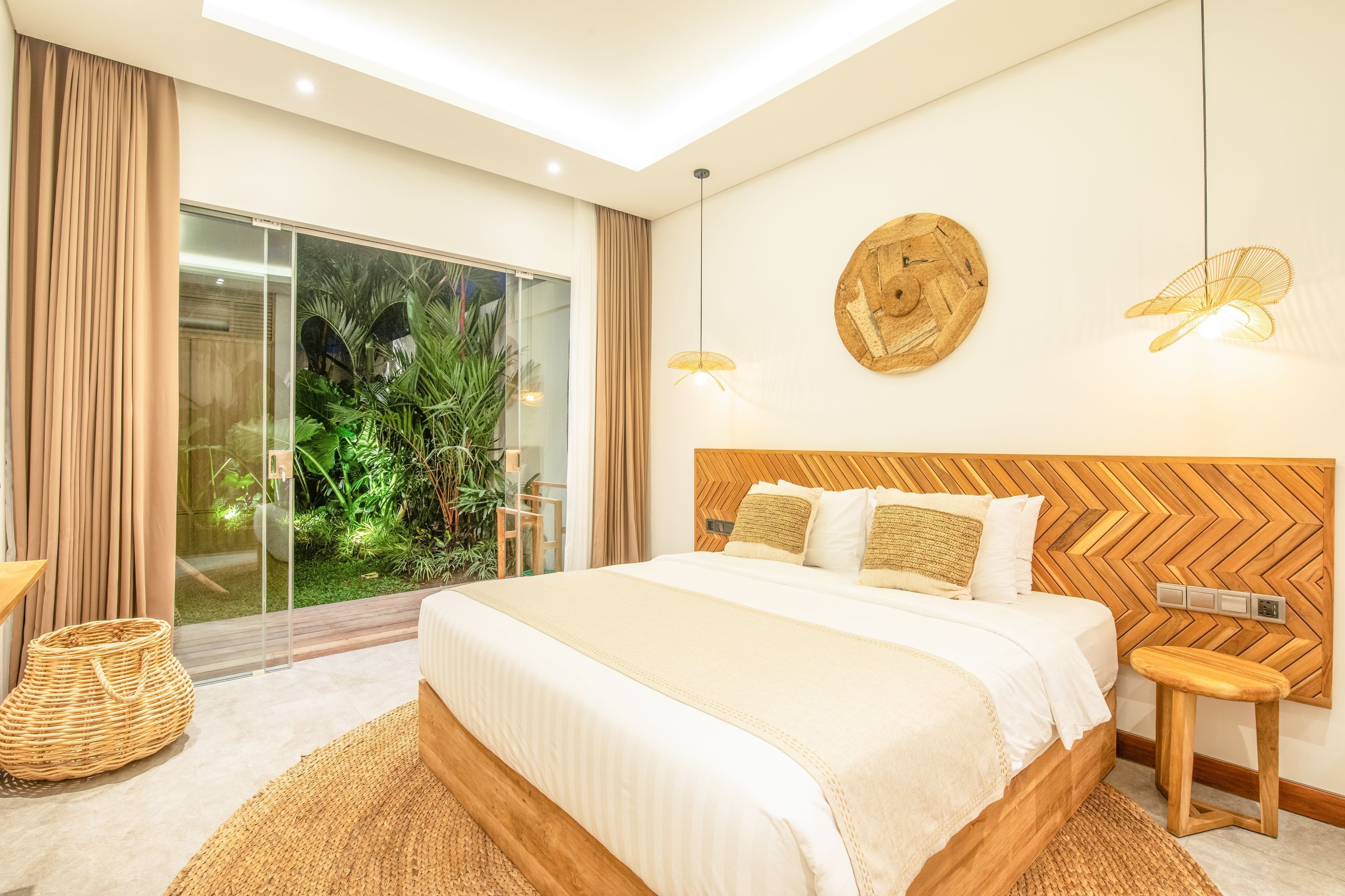 Een moderne slaapkamer met een groot bed, een houten hoofdbord, een klein krukje en uitzicht op groen via een glazen deur.